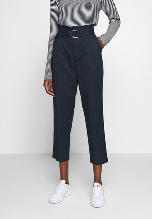 Personne portant un pantalon court taille haute bleu marine avec ceinture et baskets blanches, associé à un top rayé noir et blanc à manches longues.