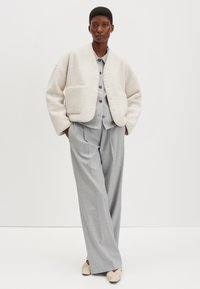 Giacca color crema, di tessuto strutturato, con grande tasca frontale; sotto un abito grigio con pantaloni a gamba larga; scarpe basse beige; sfondo semplice.