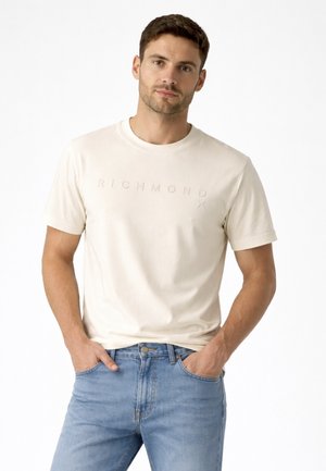 Uomo con capelli corti castani che indossa una t-shirt beige con il logo Richmond e jeans blu, con le mani nelle tasche, su sfondo bianco.