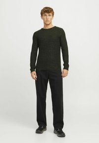 Dunkelgrüner Strickpullover mit langen Ärmeln, Rundhalsausschnitt und strukturiertem Stoff. Kombiniert mit schwarzen weit geschnittenen Hosen und schwarzen Schuhen.