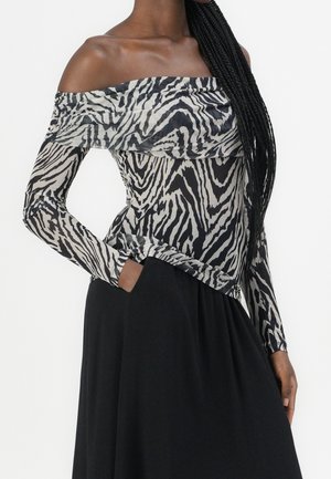 Kvinde med langt flettet hår iført en sort og hvid zebra-print off-shoulder top og et sort skørt med den ene hånd i lommen.