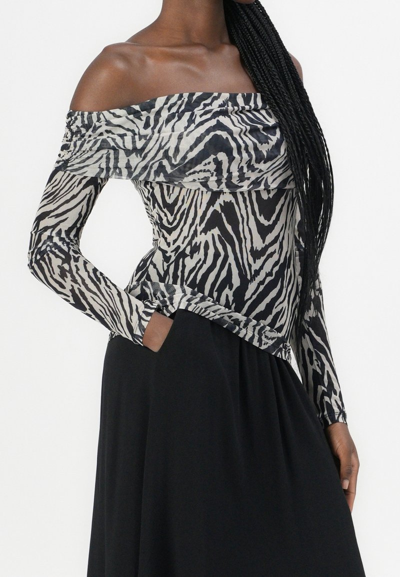 Femme aux longs cheveux tressés portant un top épaules dénudées à imprimé zèbre noir et blanc et une jupe noire, une main dans la poche.
