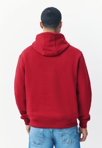 Rode hoodie met een grote capuchon aan de achterkant, geribbelde manchetten en zoom. Gladgeweven stof. Gecombineerd met lichtblauwe spijkerbroek.