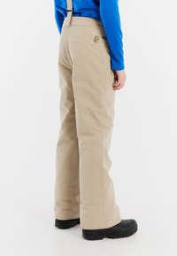 Beige skibroek met verstelbare banden, vervaardigd uit waterdicht materiaal. Voorzien van zwarte laarzen en een subtiel logo op de achterkant.