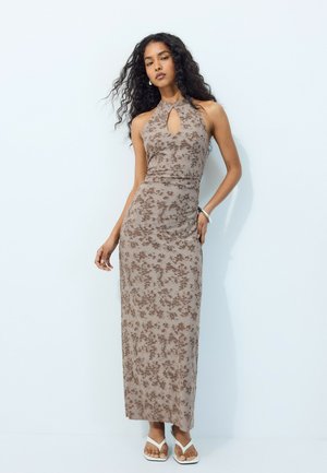 Femme aux longs cheveux bouclés portant une robe maxi marron fleurie sans manches à col halter et des sandales blanches, debout contre un mur blanc.