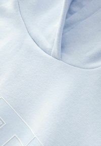 Svetlo modrá mikina s kapucňou vyrobená z mäkkého materiálu. Obsahuje textúrovaný povrch a malú, obrysovú grafiku v ľavom dolnom rohu.