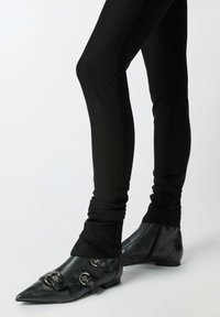 Crne uske tajice u kombinaciji s crnim čizmama do gležnja s šiljastim vrhom, koje imaju srebrne kopče i teksturiranu, sjajnu površinu.