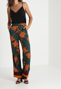 Camisola nera con scollatura smerlata abbinata a pantaloni a gamba larga caratterizzati da un motivo floreale verde e pois, con polsini arancioni a contrasto.