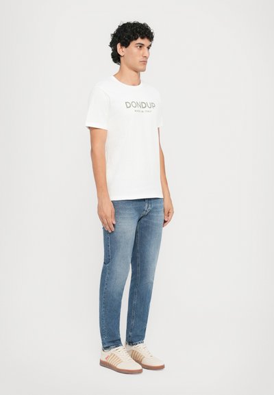 White cotton t-shirt with "DONDUP MADE IN ITALY" printed in gray, paired with faded blue jeans and beige sneakers with red accents.