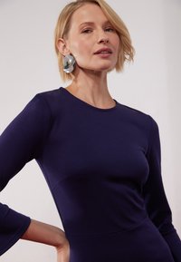 Rochie ajustată de culoare bleumarin, cu mâneci lungi și gât rotund. Prezintă o textură netedă și detalii subtile la cusături. Cercei mari, circulare, din argint.
