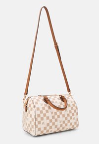 Ruskeakeltainen ja kerma ruudullinen käsilaukku, jossa kukka-aiheiset koristeet, nahkahihnat ja säädettävä olkahihna. Laukulla on pyöreä muoto ja kullanväriset metalliosat.