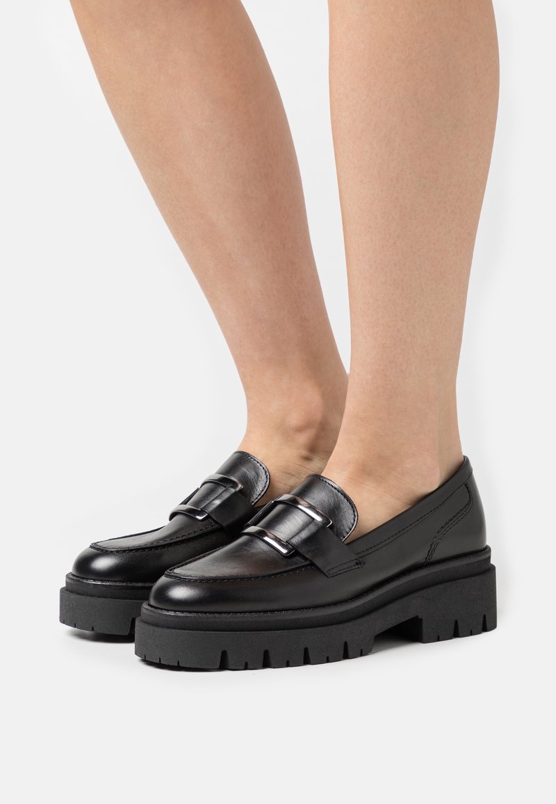 Mocassins en cuir noir avec une semelle en caoutchouc noir épaisse et un détail de boucle métallique. Texture lisse avec un design à bout arrondi et des lignes élégantes.