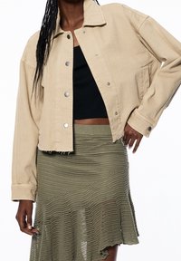 Beige spijkerjack met een cropped ontwerp, zilveren drukknopen en een rafelige zoom. Gecombineerd met een zwart topje en een groene gebreide uitlopende rok.