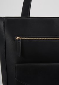 Sac fourre-tout en cuir noir avec une texture lisse, doté d'une fermeture éclair dorée et d'une petite tirette, ainsi qu'une poche avant avec rabat.