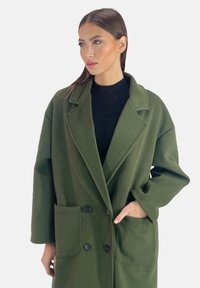 Abrigo largo verde oliva de tela suave, con cuello muesca, botones dobles y dos bolsillos delanteros.