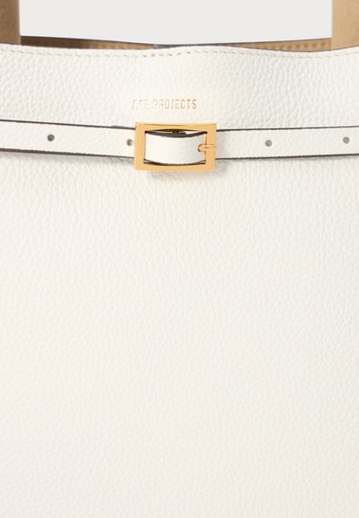 Bolso tote de cuero blanco con una superficie texturizada, un hebilla de metal dorado y costuras y acentos sutiles. El nombre de la marca está grabado en el lateral.