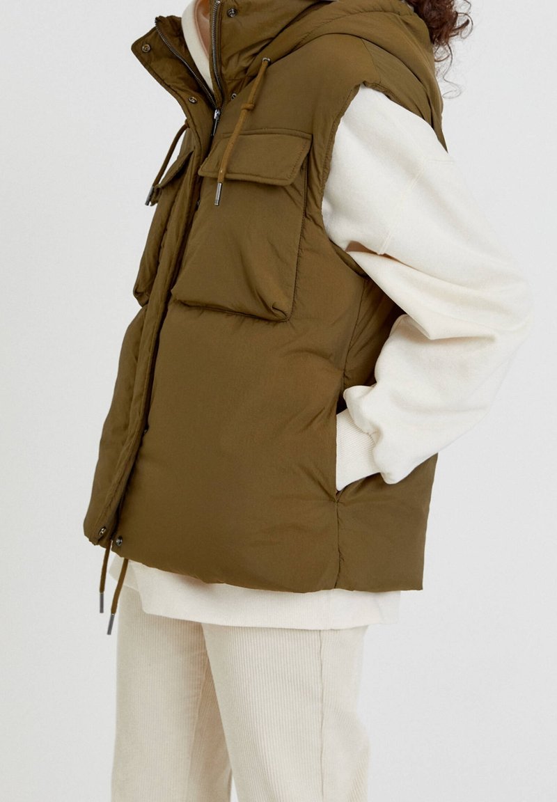 Olivgrüne, gepolsterte Weste mit Kapuze, ausgestattet mit zwei Fronttaschen und einer Kordelzugdetail. Trägt man über einem weißen Pullover und beigen Cordhosen.