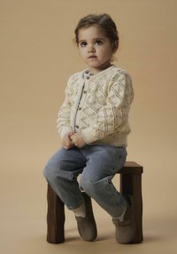 Stickad krämfärgad cardigan med diamantmönster, träknappar och ribbat nederkant; ljusblå jeans med trasiga mudd, grå mjuka tofflor. Brun träpall.