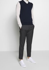 Marinblå ärmlös tröja över en ljus polotröja, parat med mörka byxor och vita sneakers; släta texturer och kontrasterande färger.