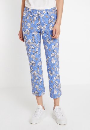 Kvinde iført korte blå bukser med hvidt og brunt blomsterprint og hvide sneakers, stående mod en ensfarvet hvid baggrund.