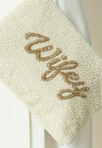 Pochette blanche perlée avec broderie de perles dorées épelant « Mrs. » sur le devant, tenue devant un fond blanc et beige.