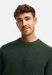 Suéter de punto acanalado verde oscuro con cuello redondo y textura suave, que presenta ligeras variaciones en color y profundidad.