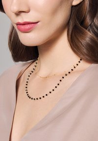Wielowarstwowe złote naszyjniki z czarnymi koralikami i delikatnym wzorem łańcuszka, spoczywające na miękkim, jasnym materiale.