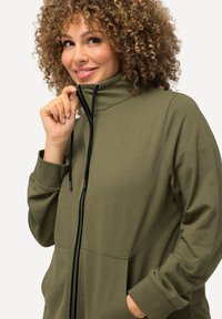 Chaqueta de cremallera verde oliva con un alto cuello, bolsillos frontales, cordones negros y una textura suave.