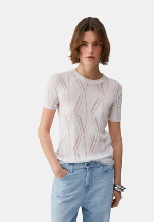 Giovane donna con capelli corti castani che indossa una maglietta lavorata a maglia bianca a maniche corte con motivo e jeans azzurri, in piedi con una mano in tasca.