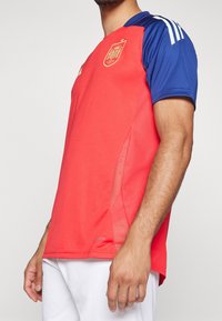 Červený fotbalový dres s modrým límcem a rukávy, zdobený bílými pruhy adidas. Zlatá embléma na hrudi, síťovaná boční panel pro prodyšnost.