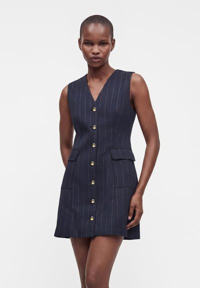 Femme aux cheveux courts portant une robe sans manches bleu marine à fines rayures avec des boutons dorés et des poches à rabat, debout devant un fond blanc uni.