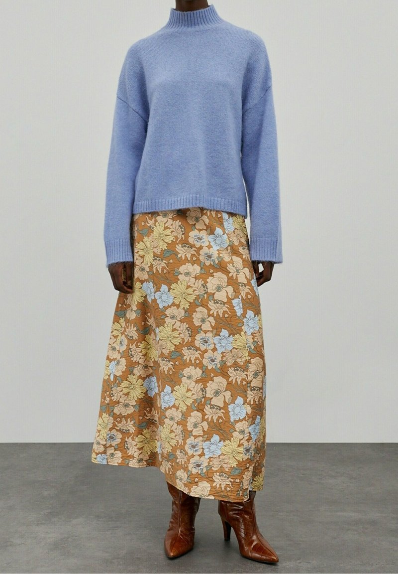 Personne portant un pull bleu clair, une jupe longue fleurie marron et des bottes marron pointues à talons hauts, debout sur un sol gris.