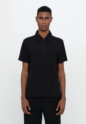 Czarna koszulka polo z krótkim rękawem i zamkiem błyskawicznym przy kołnierzyku, charakteryzująca się gładką teksturą i luźnym krojem, noszona na czarnych spodniach.