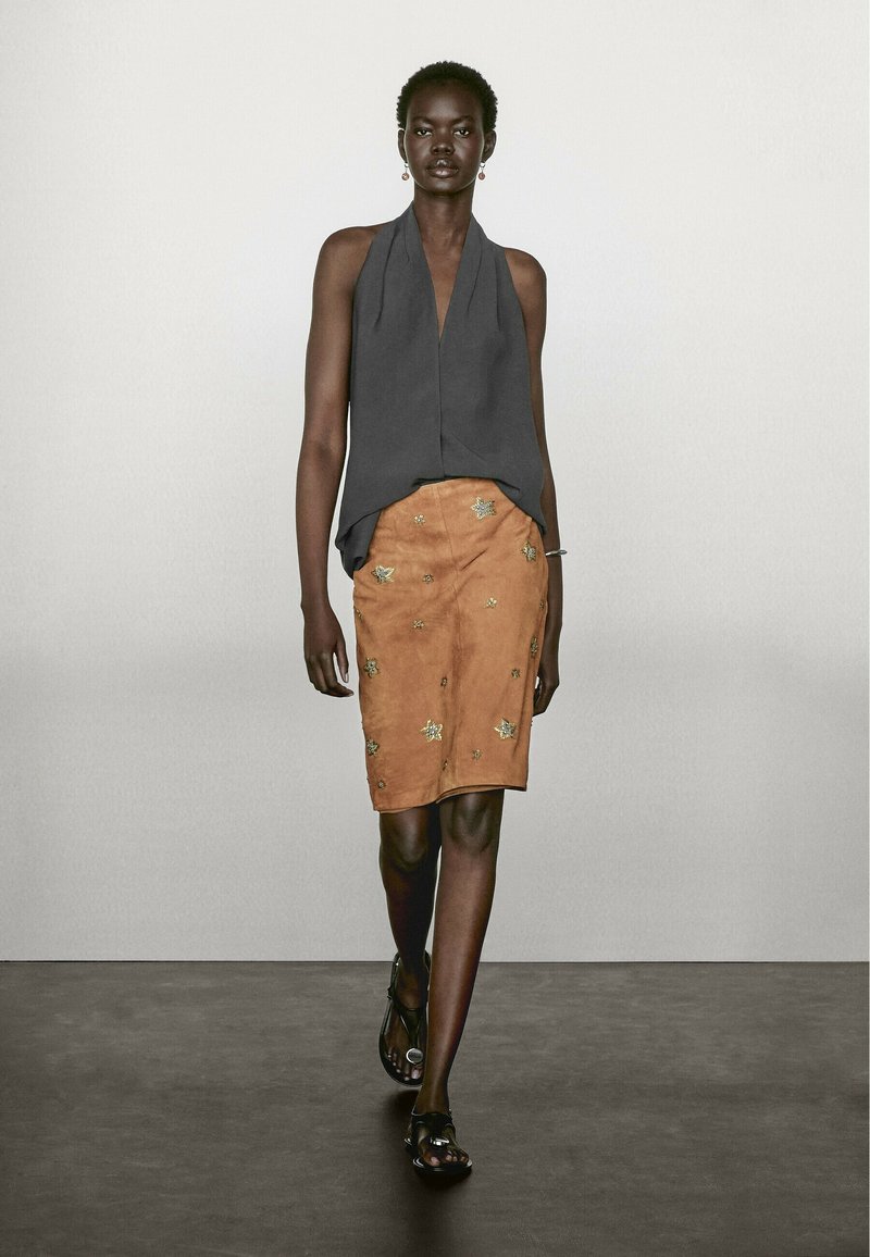 Ärmelloses graues Oberteil, A-Linien-Design, kombiniert mit einem beige-suede Rock, der goldene gestickte Blattmuster und schwarze Sandalen aufweist.