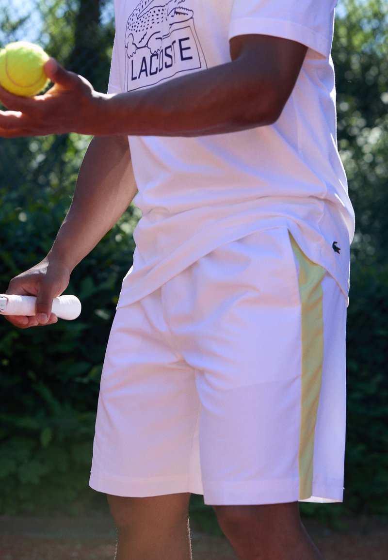 Person som bär en vit Lacoste tennisoutfit, håller en tennisboll i ena handen och racketens handtag i den andra, utomhus.