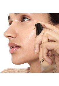 Personne appliquant un fond de teint liquide beige sur la joue à l'aide d'un flacon pompe noir, mettant en valeur une peau lisse, éclatante et la texture du maquillage.