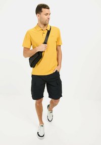 Żółta koszulka polo z krótkim rękawem, teksturowana tkanina, w połączeniu z czarnymi szortami cargo i białymi sneakersami. Nosi czarną torbę na ramię.