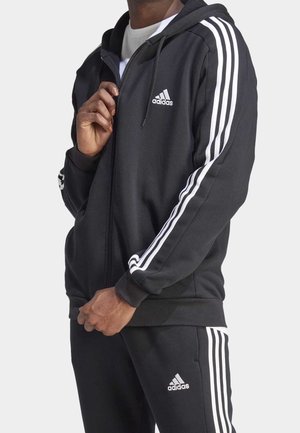 Person wearing black Adidas tracksuit with white stripes on sleeves and pants, adjusting zipper on the jacket.