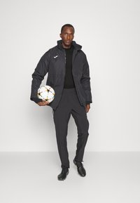 Schwarze wasserdichte Jacke mit Kapuze, schwarzes T-Shirt und dunkle Hose. Hält einen mehrfarbigen Fußball. Schwarze Fußballschuhe mit blauen Akzenten.