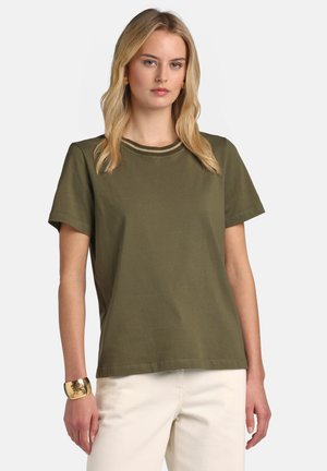 Szőke nő olívazöld rövid ujjú felsőt visel csíkos gallérral, fehér nadrágot és arany mandzsettakarkötőt a bal csuklóján.