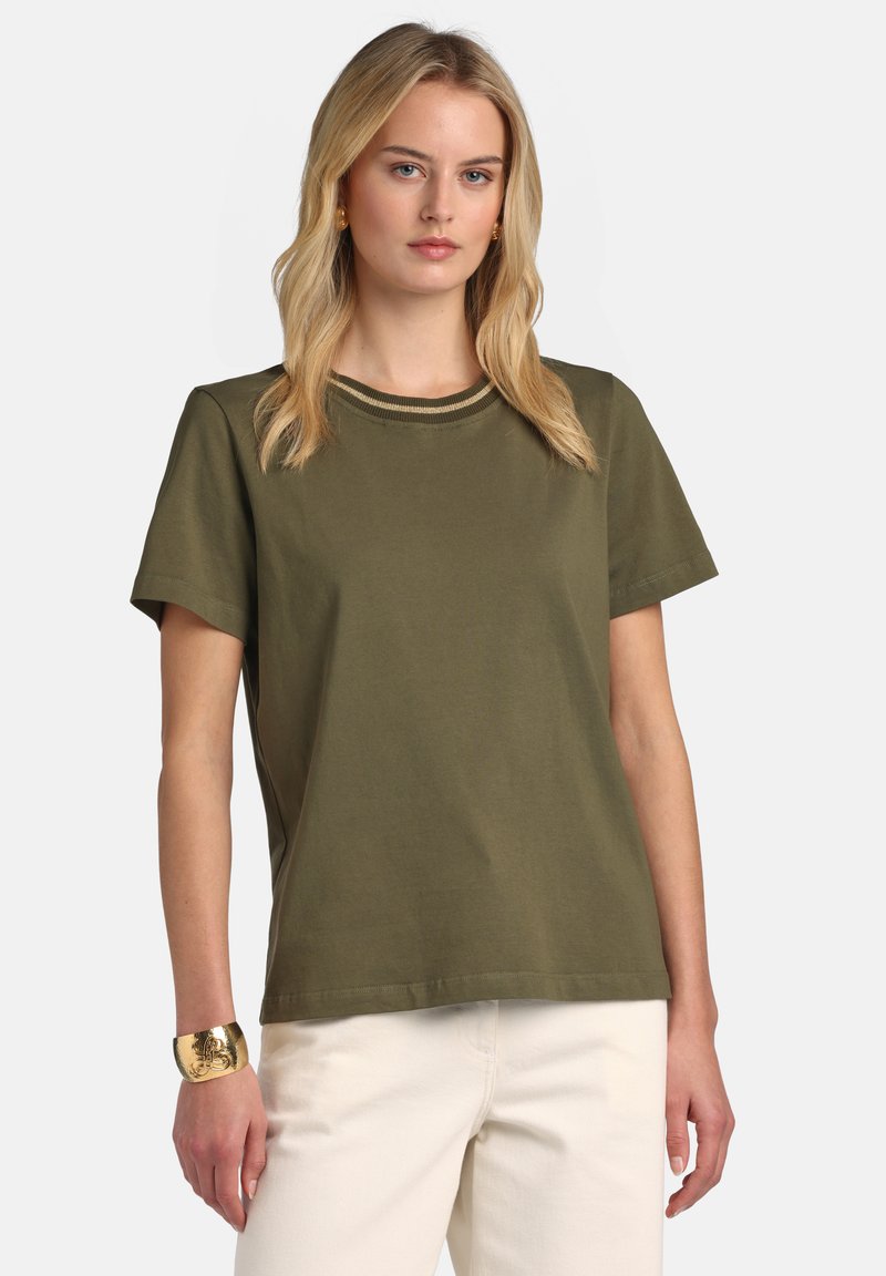 Femme blonde portant un haut à manches courtes vert olive avec col rayé, pantalon blanc et un bracelet manchette en or au poignet gauche.
