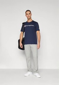 Tmavě modré bavlněné tričko s bílým logem "Champion", doplněné světle šedými tepláky a bílými teniskami. Model drží černou tašku.