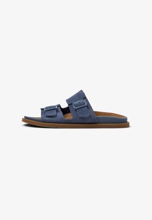 Sandale mule en daim bleu foncé, avec deux boucles de sangle réglables et une semelle plate en caoutchouc marron. Design à bout ouvert avec une texture douce.