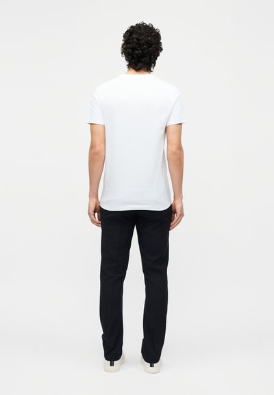 Άνδρας με μπούκλες, που στέκεται πλάτη, φοράει λευκό κοντομάνικο μπλουζάκι, μαύρο παντελόνι και λευκά αθλητικά με μαύρες ρίγες.