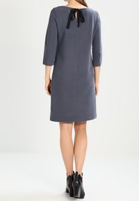 Harmaa polvipituinen mekko, jossa on kolmanneksen mittaiset hihat, takana avaus, joka on sidottu mustalla nauhalla, yhdistetty mustiin korkokenkiin.