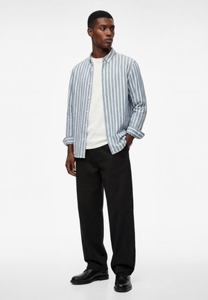 Mann steht und trägt ein blau-weiß vertikal gestreiftes Hemd über einem weißen T-Shirt, schwarze Hose und schwarze Schuhe, Hände in den Taschen.