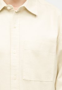 Κοντινό πλάνο ατόμου που φοράει πουκάμισο εκρού με κουμπιά, με τσέπη στο στήθος και κανονικό γιακά.