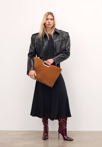 Femme portant une veste en cuir noire, une longue robe noire, et des bottes à talons hauts bordeaux, tenant un grand sac à main en daim marron avec une poignée dorée.