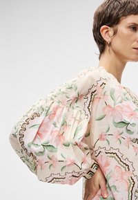 Λουλουδάτη μπλούζα με φουσκωτά μανίκια, κατασκευασμένη από ελαφρύ ύφασμα. Διαθέτει ροζ λουλούδια, πράσινα φύλλα και διακοσμητικές καφέ περιγράμματα.