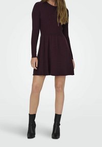 Femme portant une robe en tricot à manches longues de couleur violet foncé avec une jupe évasée et des bottines noires, debout devant un fond uni.
